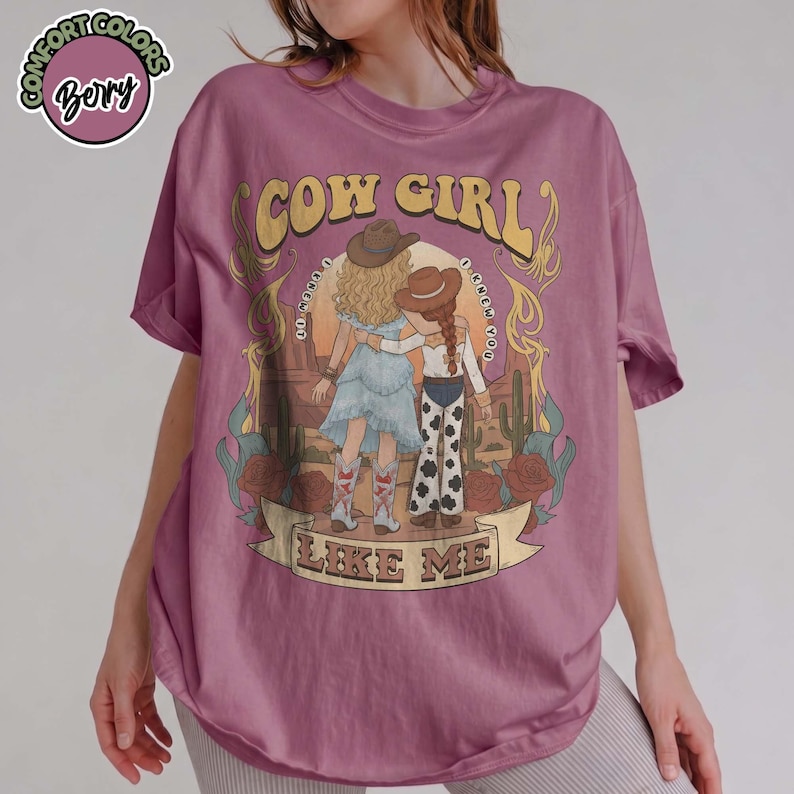 May include: A berry-colored t-shirt featuring a graphic of two figures in cowboy hats standing in a desert landscape. The text reads COW GIRL LIKE ME. The design includes cacti, roses, and decorative scrollwork in muted tones on the front of the shirt.