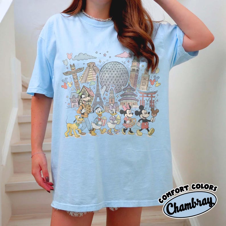 May include: Light blue t-shirt featuring a colorful graphic of Disney characters and world landmarks. The design includes Mickey Mouse, Donald Duck, and other characters. The shirt has the text "COMFORT COLORS Chambray" in a circular design.