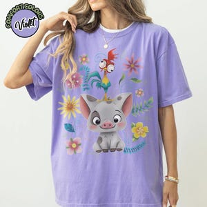 May include: Violet Comfort Colors t-shirt featuring a cartoon pig with a rooster on its head, surrounded by colorful flowers and foliage. The shirt has a relaxed fit and is a light purple color.