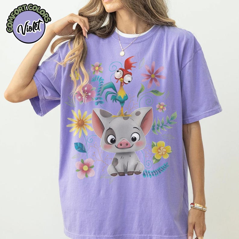 May include: Violet Comfort Colors t-shirt featuring a cartoon pig with a rooster on its head, surrounded by colorful flowers and foliage. The shirt has a relaxed fit and is a light purple color.