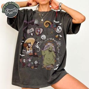 May include: Dark gray t-shirt featuring a colorful graphic design with characters from "The Nightmare Before Christmas." The design includes Jack Skellington, Sally, Oogie Boogie, and other Halloween-themed elements. The shirt has the text "NIGHTMARE" in a circular pattern.