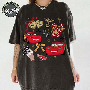 Puede incluir: Camiseta gris oscuro con un dise&ntilde;o gr&aacute;fico de los personajes de la pel&iacute;cula Cars. El dise&ntilde;o incluye a Rayo McQueen, corazones, lazos y el texto "Cars". La camiseta tiene un aspecto vintage y desgastado.