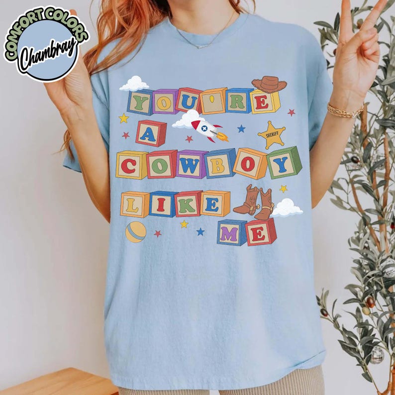 May include: A light blue t-shirt featuring a graphic print of colorful alphabet blocks spelling out the text YOU'RE A COWBOY LIKE ME. The design includes illustrations of a rocket, a brown cowboy hat, a sheriff star badge, and cowboy boots.