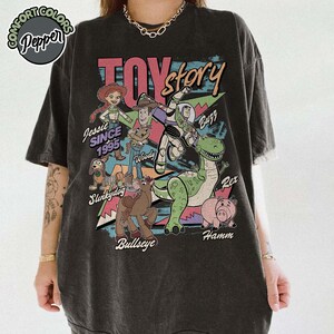 May include: Dark gray t-shirt featuring a colorful graphic of Toy Story characters, including Woody, Buzz Lightyear, and Rex. The text "Toy Story" is displayed above the characters, with "Jessie Since 1995" below.