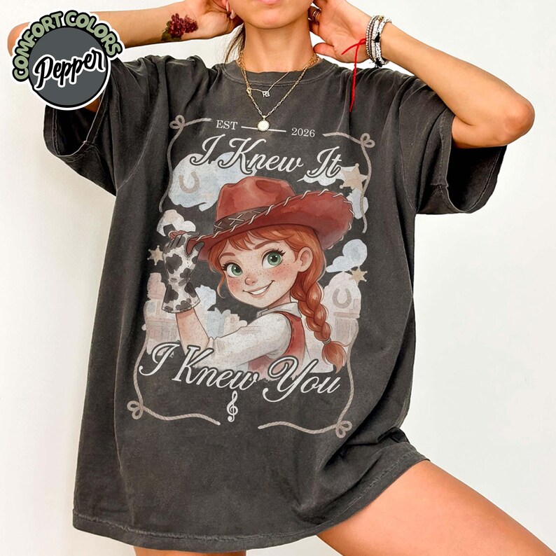 Peut inclure: Un t-shirt gris anthracite &agrave; manches courtes avec un motif de cow-girl dessin anim&eacute; aux cheveux roux et au chapeau marron. Le texte indique "EST 2026", "I Knew It" et "I Knew You". Le design inclut une bordure lasso, des &eacute;toiles et des fers.