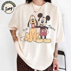May include: Ivory-colored t-shirt featuring Pluto and Mickey Mouse. Pluto is depicted in a sitting position with his tongue out, while Mickey Mouse stands beside him. The text "Pluto" and "Mickey Mouse" are also visible.