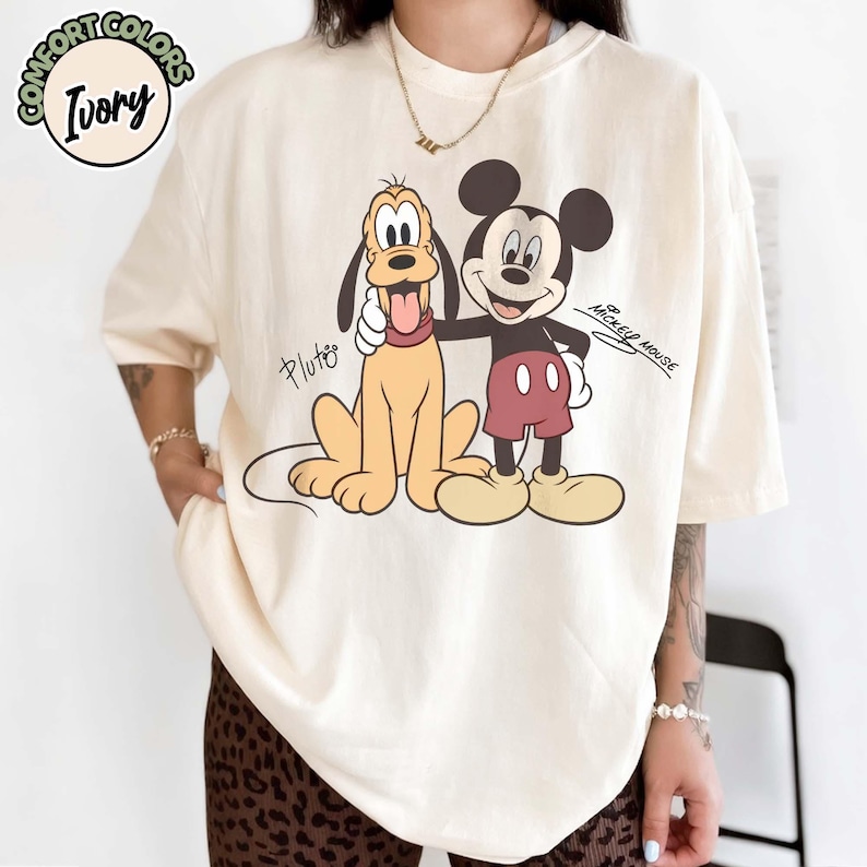 May include: Ivory-colored t-shirt featuring Pluto and Mickey Mouse. Pluto is depicted in a sitting position with his tongue out, while Mickey Mouse stands beside him. The text "Pluto" and "Mickey Mouse" are also visible.