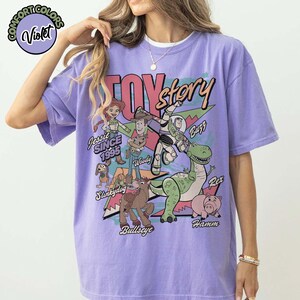 May include: A violet Comfort Colors t-shirt featuring a graphic of Toy Story characters. The design includes Jessie, Woody, Buzz, Rex, Hamm, Slinkydog, and Bullseye, with the text "TOY STORY" and "SINCE 1995".