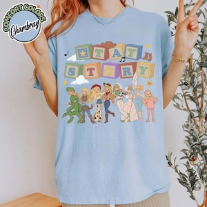 May include: A light blue t-shirt featuring a graphic print of colourful alphabet blocks spelling out the text "STAY STORY" above a line of illustrated characters in various outfits, including a green dinosaur costume and a pink dress.