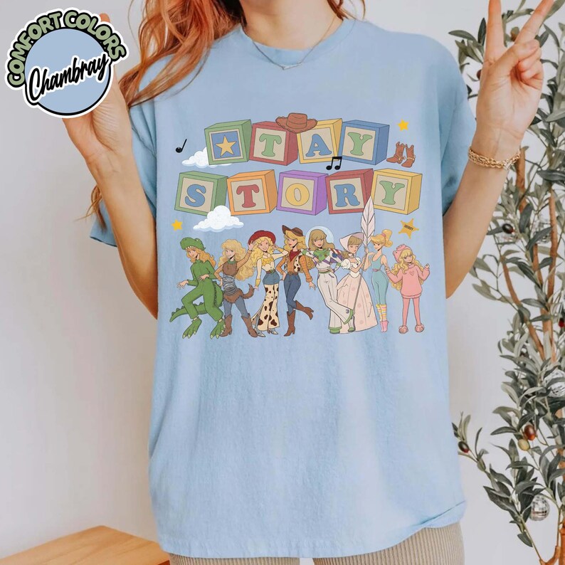 May include: A light blue t-shirt featuring a graphic print of colourful alphabet blocks spelling out the text "STAY STORY" above a line of illustrated characters in various outfits, including a green dinosaur costume and a pink dress.