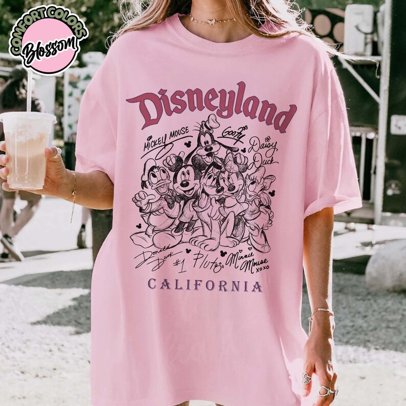 Puede incluir: Camiseta rosa claro con un gr&aacute;fico de Disneyland que presenta a Mickey Mouse, Goofy, Donald Duck, Pluto, Minnie Mouse y Daisy Duck. El gr&aacute;fico incluye la palabra "Disneyland" y "California".