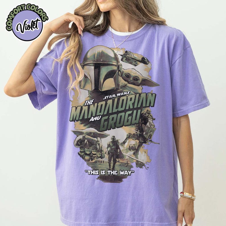 May include: A violet short-sleeve t-shirt featuring a graphic print of The Mandalorian and Grogu. The design includes the text "The Mandalorian and Grogu" and "This is the way" along with various Star Wars character illustrations in muted tones.