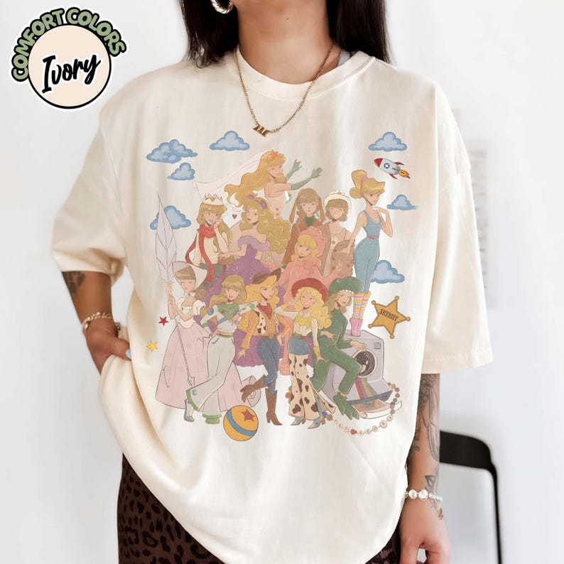 May include: An ivory t-shirt featuring a graphic print of multiple animated characters in various outfits, including a space suit, cowgirl attire, and dresses. The design includes blue clouds, a small rocket, a star, and a ball. Text reads COMFORT COLORS IVORY.