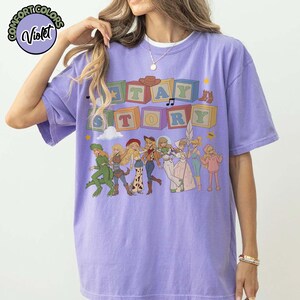 May include: A light purple short-sleeve t-shirt featuring a graphic print of colourful alphabet blocks spelling STAY STORY above a line of illustrated characters in various outfits. A circular logo in the top left corner reads COMFORT COLORS Violet.