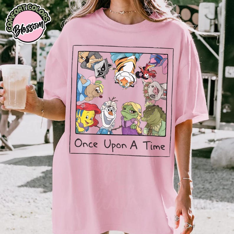 Puede incluir: Camiseta rosa claro con un dise&ntilde;o enmarcado de personajes de dibujos animados y el texto "Once Upon A Time". El dise&ntilde;o incluye un mapache, un tigre y otros personajes animados. La camiseta tiene mangas cortas y un corte holgado.