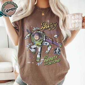 May include: Brown t-shirt featuring a graphic of Buzz Lightyear from Toy Story. The design includes the text "Buzz Lightyear" and "To Infinity and Beyond" in a vintage style. The shirt has a crew neck and short sleeves.