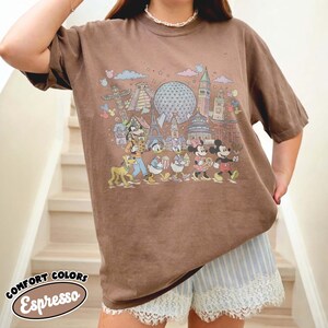 May include: Brown Comfort Colors Espresso t-shirt featuring a colorful graphic of Disney characters and landmarks. The shirt has a relaxed fit and short sleeves. The text "COMFORT COLORS Espresso" is printed on the bottom left corner.