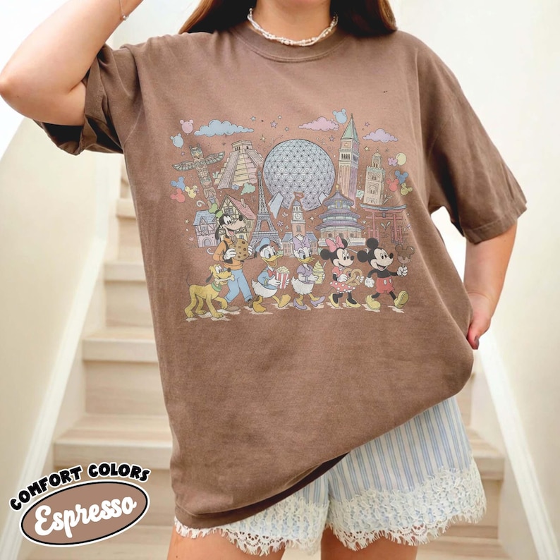 May include: Brown Comfort Colors Espresso t-shirt featuring a colorful graphic of Disney characters and landmarks. The shirt has a relaxed fit and short sleeves. The text "COMFORT COLORS Espresso" is printed on the bottom left corner.