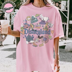 Puede incluir: Camiseta rosa claro con un gr&aacute;fico colorido de Disneyland. El dise&ntilde;o incluye ilustraciones de un castillo, una noria y otros elementos ic&oacute;nicos del parque, con la palabra "Disneyland" en una fuente l&uacute;dica.