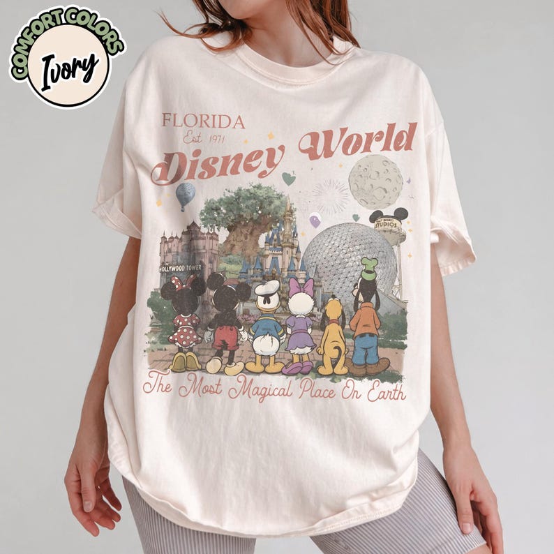 May include: Ivory-colored t-shirt with a Disney World graphic. The design features Disney characters in front of park landmarks, with text that reads "Florida Est. 1971 Disney World" and "The Most Magical Place On Earth."