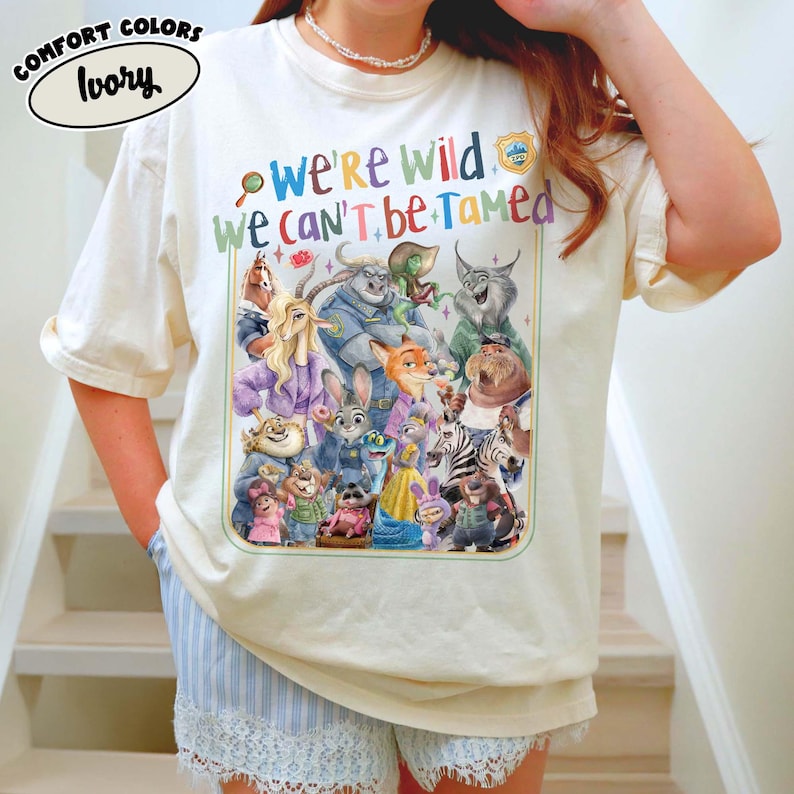 May include: Ivory-colored t-shirt with a colorful graphic featuring characters from Zootopia and the text "We're Wild We Can't Be Tamed." The shirt is a Comfort Colors brand.