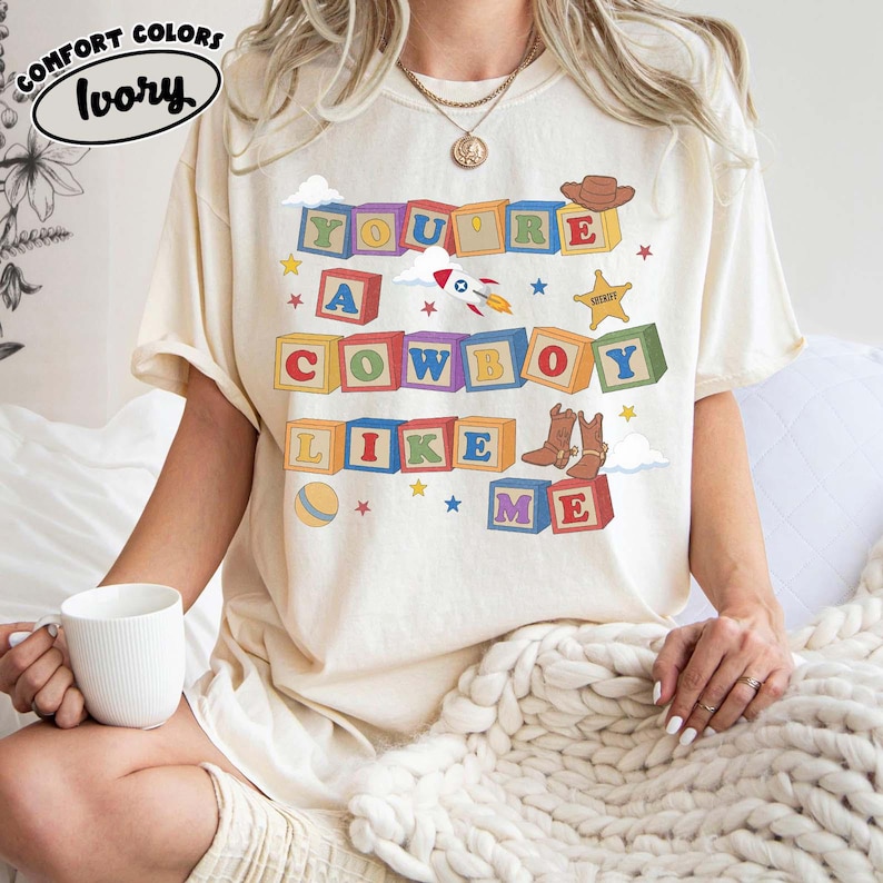 May include: An ivory short-sleeve t-shirt featuring a graphic of colorful alphabet blocks spelling out the text You're A Cowboy Like Me. The design includes illustrations of a brown cowboy hat, a rocket ship, a sheriff star, and cowboy boots.
