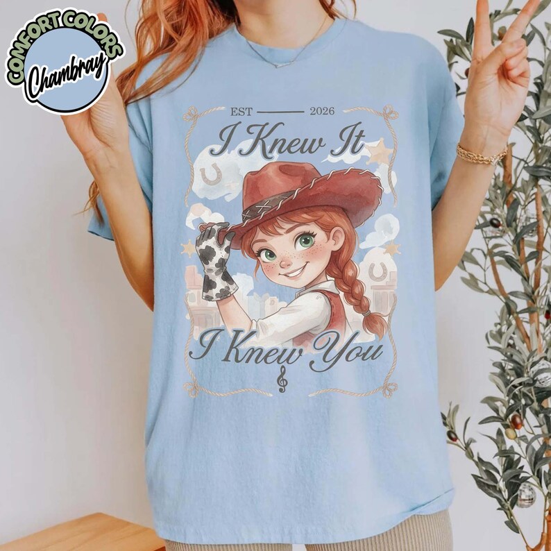 Peut inclure: Un t-shirt bleu clair avec une illustration de cow-girl portant un chapeau marron et un gant &agrave; motif de vache. Le texte indique EST 2026, I Knew It et I Knew You, accompagn&eacute; d'une note de musique. Le motif inclut des nuages et des &eacute;toiles.