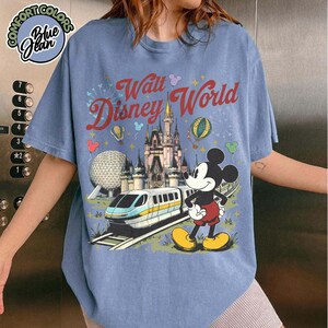 May include: A blue t-shirt featuring a Walt Disney World graphic. The design includes Cinderella's Castle, Mickey Mouse, a monorail, and the text "Walt Disney World". The shirt has a Comfort Colors label.