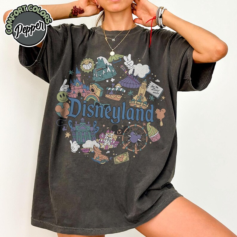 Puede incluir: Camiseta gris oscuro con un gr&aacute;fico colorido de Disneyland. El dise&ntilde;o incluye elementos ic&oacute;nicos del parque como un castillo, una noria y helado, con la palabra "Disneyland". La camiseta tiene un aspecto vintage y desgastado.