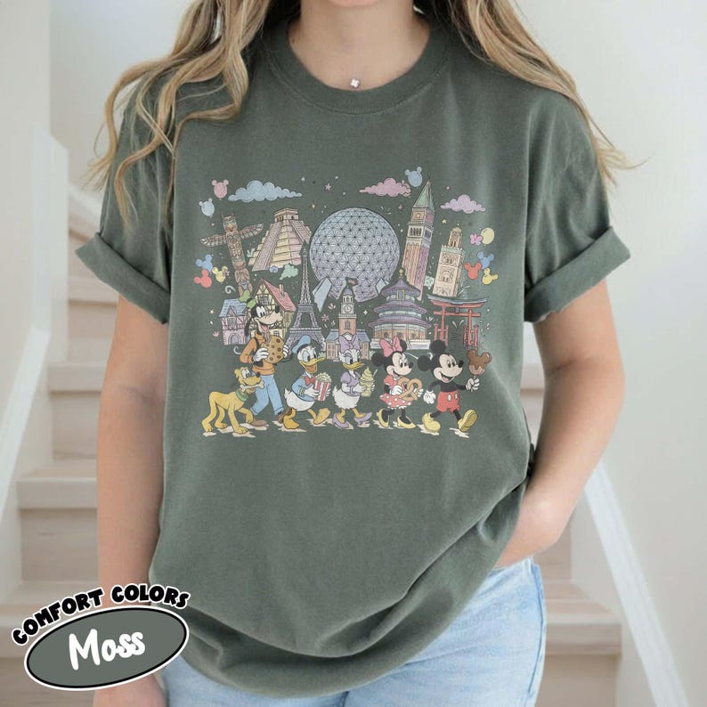 May include: Moss green t-shirt featuring a colorful graphic of Disney characters and landmarks from around the world. The design includes Mickey Mouse, Minnie Mouse, Donald Duck, and Goofy. The shirt has the words "COMFORT COLORS" and "Moss" printed on it.