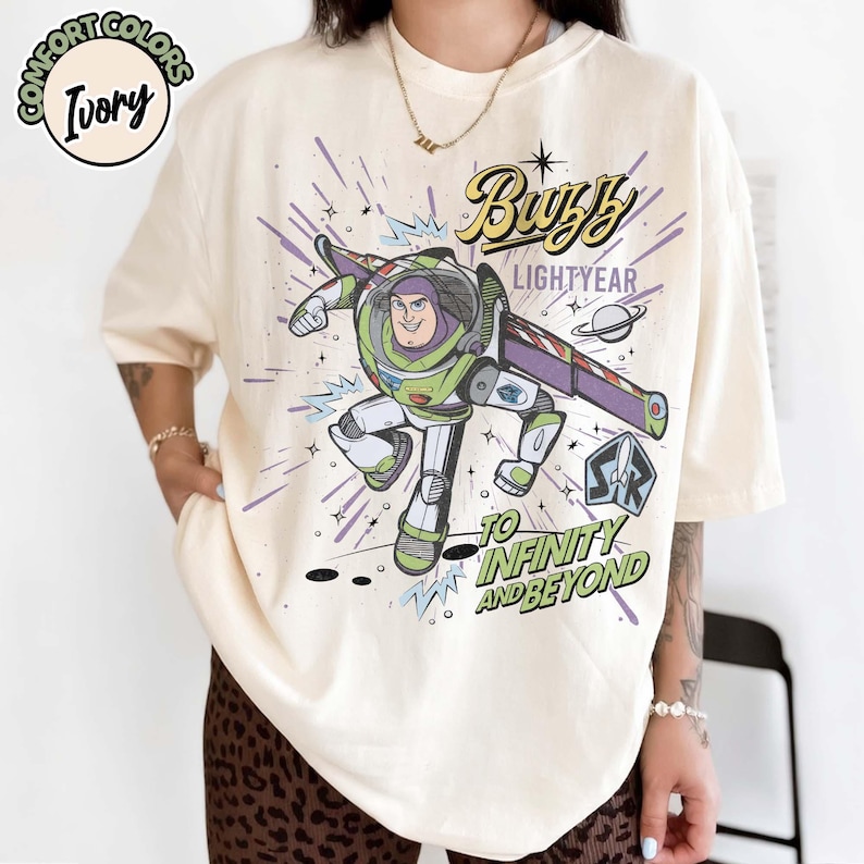 May include: Ivory-colored t-shirt featuring a graphic of Buzz Lightyear in a dynamic pose, with the words "Buzz Lightyear" and "To Infinity and Beyond." The design includes a space-themed background with purple and blue accents.