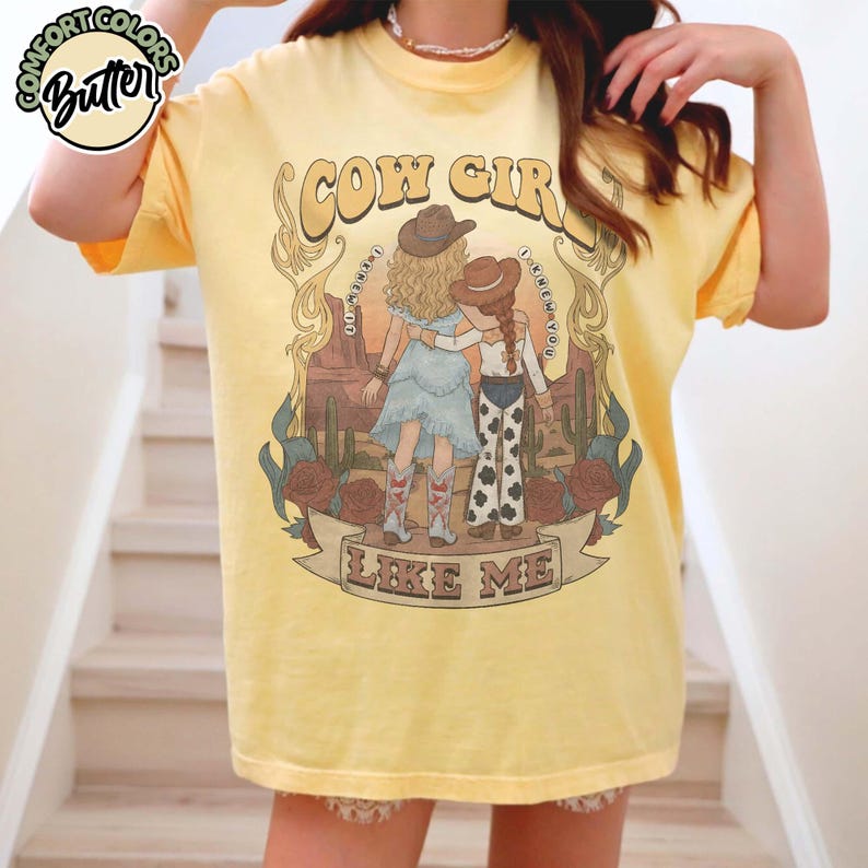 May include: A pale yellow short-sleeve t-shirt featuring a graphic print of two figures in cowboy hats standing in a desert landscape with cacti and roses. The text reads COW GIRL and LIKE ME, with the phrase I KNEW YOU I KNEW YOU arched around.