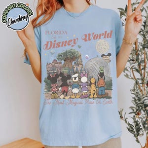 May include: Light blue t-shirt featuring a Disney World graphic. The design includes the text "Florida Est. 1971 Disney World" and a cartoon image of Disney characters in front of park landmarks. The phrase "The Most Magical Place On Earth" is also printed on the shirt.