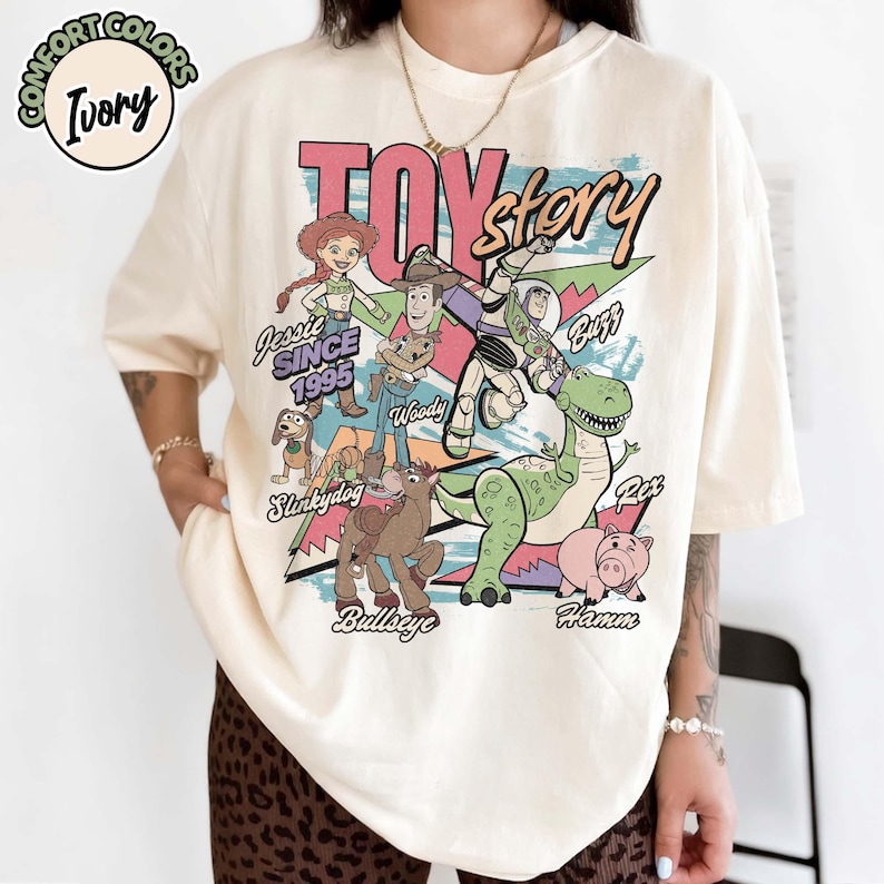 May include: Ivory-colored t-shirt featuring a colorful graphic of Toy Story characters, including Woody, Buzz Lightyear, and Rex. The design includes the text "Toy Story" and "Jessie Since 1995". The shirt has a relaxed fit.