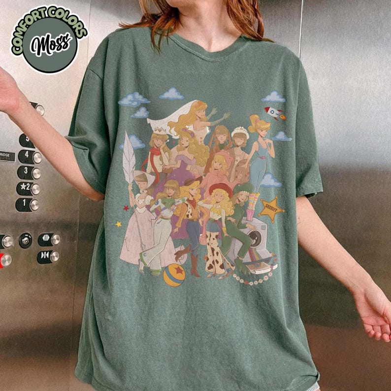 May include: A moss green short-sleeve t-shirt featuring a graphic print of various animated female characters in diverse outfits, including a cowgirl, a space ranger, and a bride. A small rocket, clouds, and a star are also printed on the shirt.