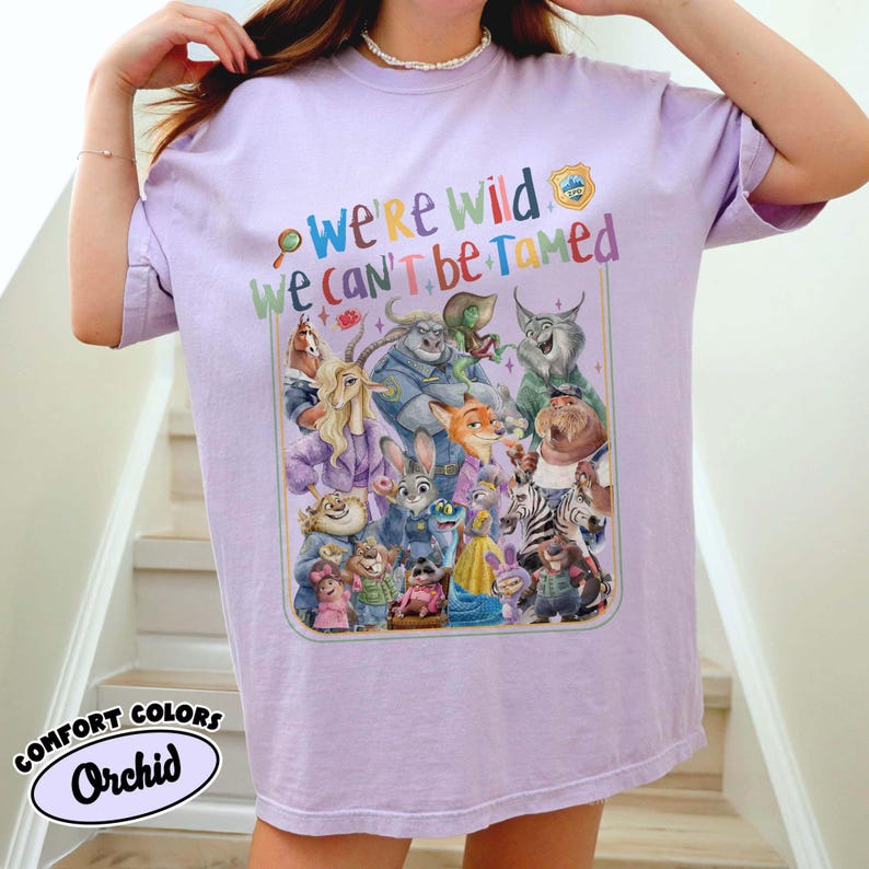 May include: A lilac-colored t-shirt with a colorful graphic featuring characters from a popular animated movie. The text "We're Wild We Can't Be Tamed" is above the graphic. The shirt also has the words "Comfort Colors" and "Orchid" printed on it.