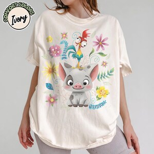 May include: Ivory-colored t-shirt featuring a cartoon pig with a rooster on its head, surrounded by colorful flowers and foliage. The shirt has the words "Comfort Colors" and "Ivory" printed on it.
