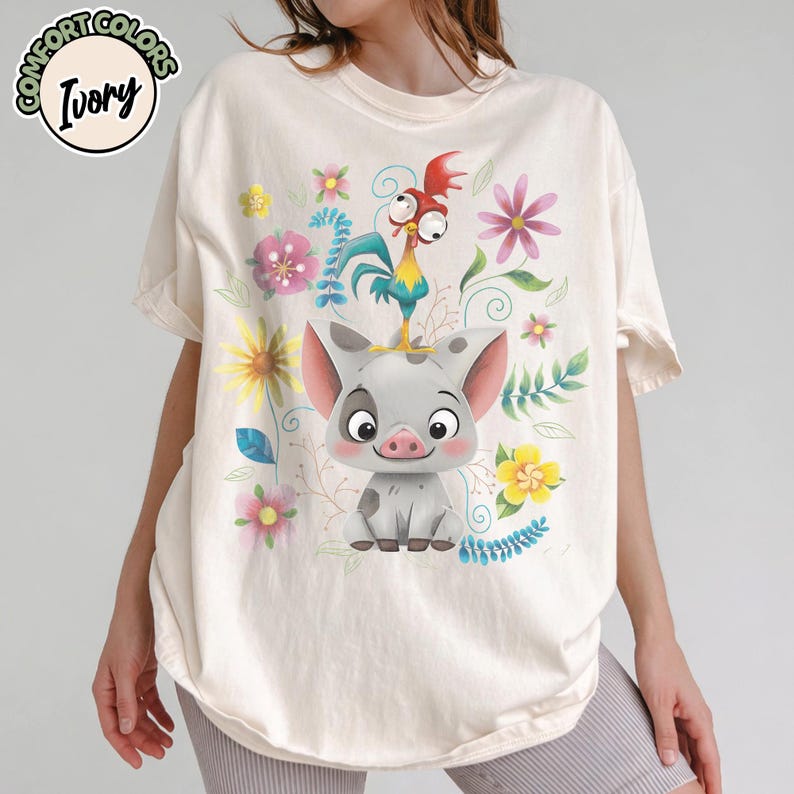 May include: Ivory-colored t-shirt featuring a cartoon pig with a rooster on its head, surrounded by colorful flowers and foliage. The shirt has the words "Comfort Colors" and "Ivory" printed on it.