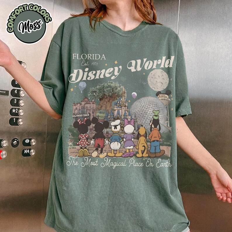 May include: Green t-shirt with a Disney World graphic. The graphic features Disney characters, the text "FLORIDA Est. 1971 Disney World", and the phrase "The Most Magical Place On Earth". The shirt has the Comfort Colors Moss logo.