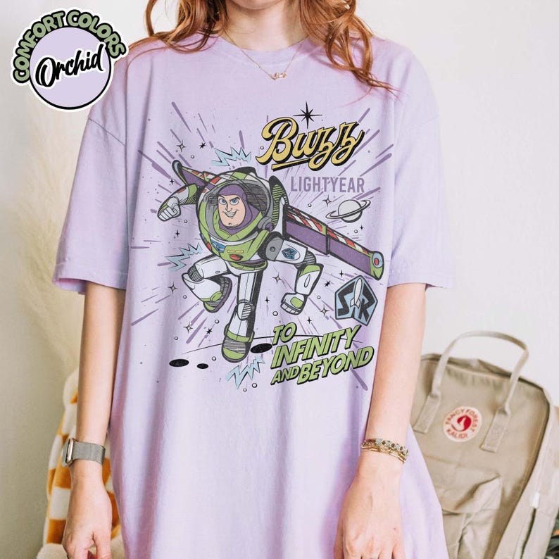 May include: A light purple t-shirt featuring a graphic of Buzz Lightyear from Toy Story. The design includes the text "Buzz Lightyear" and "To Infinity and Beyond". The shirt has short sleeves and a relaxed fit.