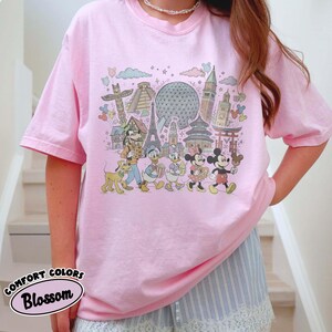 May include: Pink t-shirt featuring a colorful graphic of Disney characters and landmarks. The design includes Mickey Mouse, Minnie Mouse, Donald Duck, and Goofy, with the Eiffel Tower and Spaceship Earth. The text "COMFORT COLORS Blossom" is also visible.