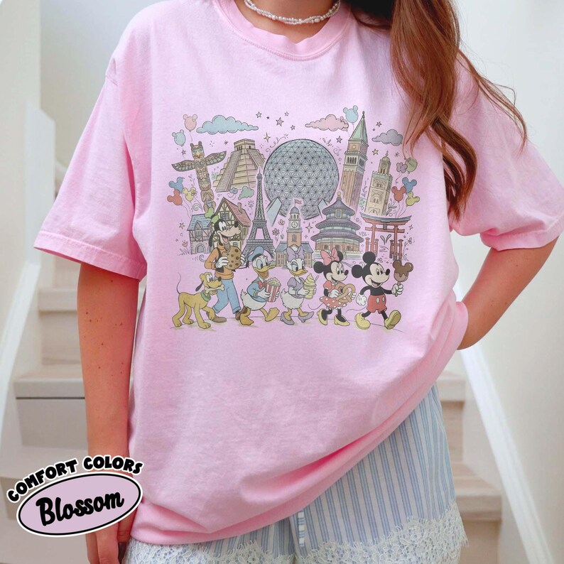 May include: Pink t-shirt featuring a colorful graphic of Disney characters and landmarks. The design includes Mickey Mouse, Minnie Mouse, Donald Duck, and Goofy, with the Eiffel Tower and Spaceship Earth. The text "COMFORT COLORS Blossom" is also visible.