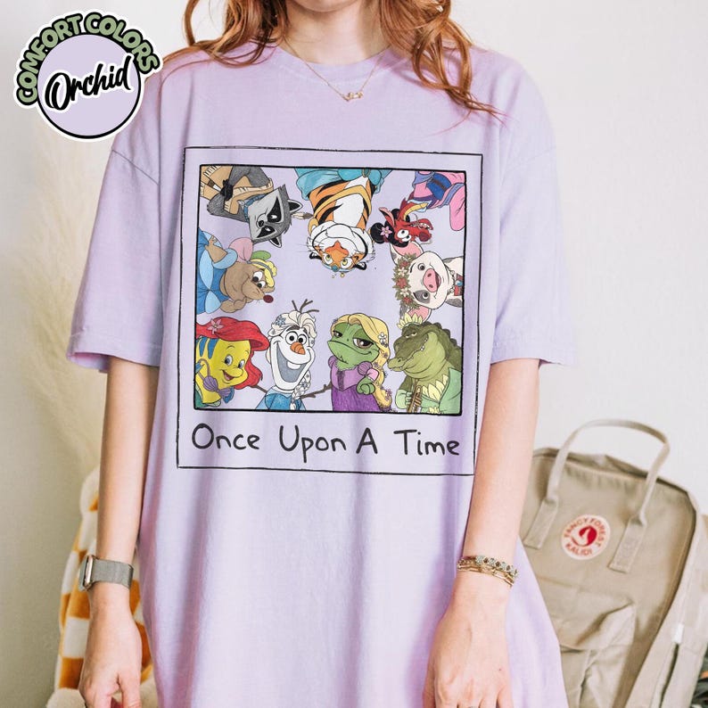 Puede incluir: Una camiseta lila con un gr&aacute;fico enmarcado de dibujos animados de personajes como un mapache, un tigre y una rana. El texto "Once Upon A Time" est&aacute; debajo. La camiseta tiene el logo Comfort Colors Orchid.