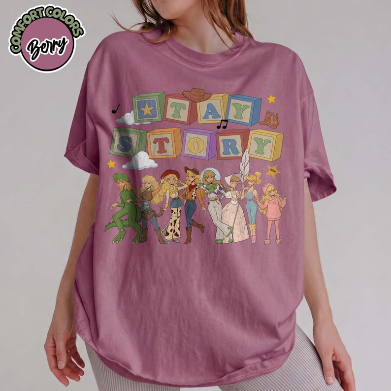 May include: A berry-coloured short-sleeve t-shirt featuring a graphic print of colourful alphabet blocks spelling TAY STORY above a line of eight illustrated characters in various outfits, including a dinosaur costume, western wear, and a gown.