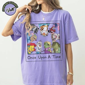 Puede incluir: Una camiseta violeta con un dise&ntilde;o enmarcado de personajes de dibujos animados, incluyendo a Ariel, Olaf y Rapunzel. El texto "Once Upon A Time" est&aacute; impreso debajo. La camiseta tiene mangas cortas y cuello redondo.