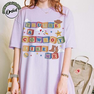 May include: A light purple t-shirt featuring a graphic of colorful alphabet blocks spelling out the phrase YOU'RE A COWBOY LIKE ME. The design includes illustrations of a rocket, a sheriff badge, a cowboy hat, cowboy boots, and a ball.