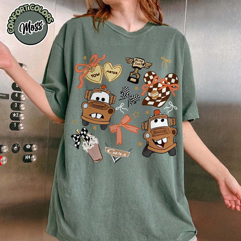 Puede incluir: Camiseta verde musgo con gr&aacute;ficos de dibujos animados de los personajes de la pel&iacute;cula Cars, Mater y Rayo McQueen. La camiseta presenta el texto "Cars" y otros elementos decorativos.