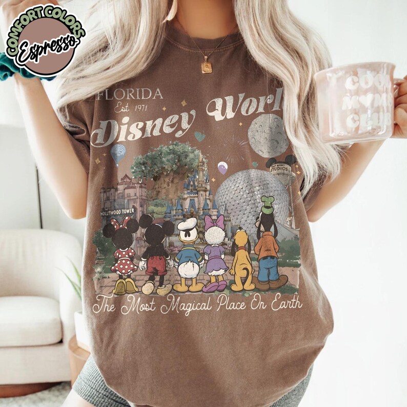 May include: Brown t-shirt featuring a Disney World graphic with Mickey and Minnie Mouse, Donald Duck, Daisy Duck, Goofy, and Pluto. The shirt has the text "Florida Est. 1971 Disney World" and "The Most Magical Place On Earth."