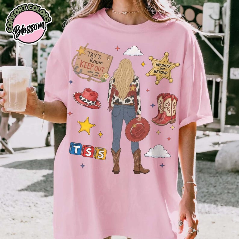 May include: A light pink t-shirt featuring a graphic of a person with long blonde hair, wearing a cowgirl outfit. The design includes a sign reading TAY'S ROOM KEEP OUT, a sheriff star with INFINITY AND BEYOND, a red hat, boots, and TS5 blocks.