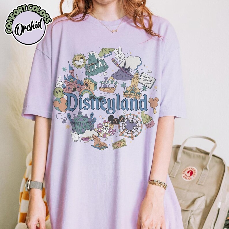 Puede incluir: Camiseta lila claro Comfort Colors con un dise&ntilde;o de Disneyland. El dise&ntilde;o incluye iconos de Disneyland, como el castillo y una noria, con la palabra "Disneyland". La camiseta tiene mangas cortas y un corte holgado.